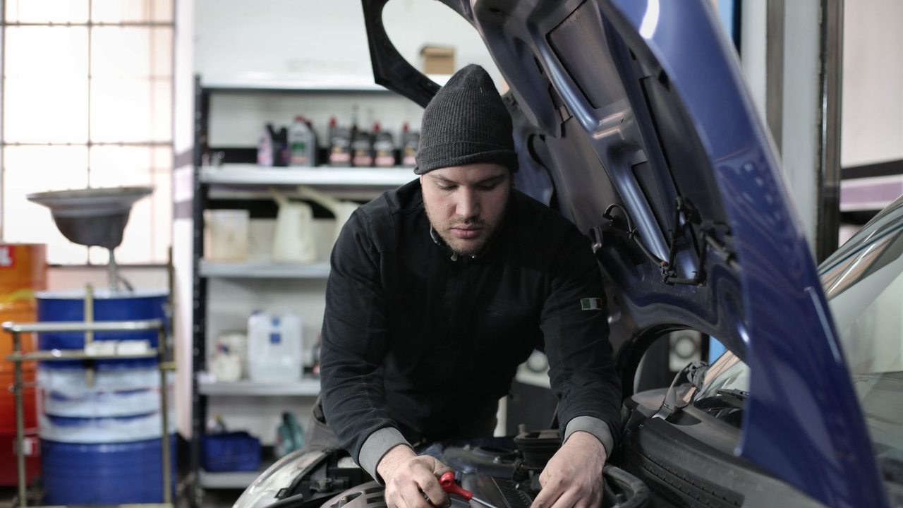 A mechanic in a black hat examines a car engine with the bonnet open in a workshop.