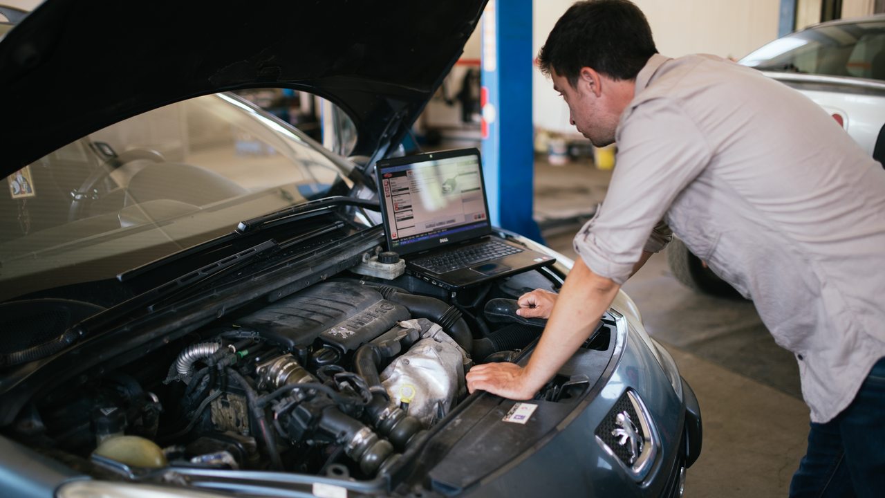 A mechanic consults a laptop beside a car engine in a workshop.