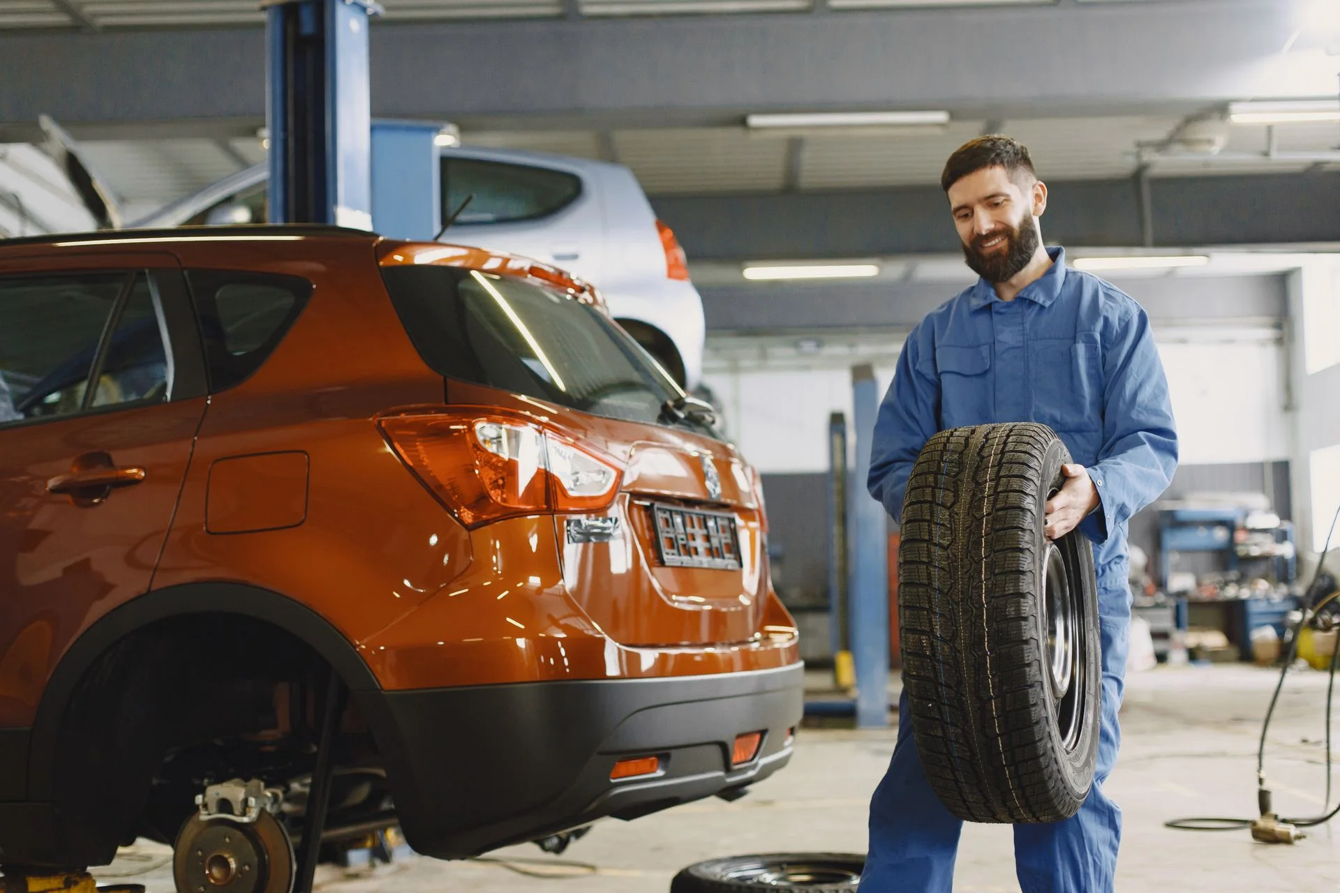 A smiling mechanic holds a tyre next to a car in his workshop.